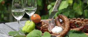 Zwei Weinglaser mit Wasser, eine grüne Paprika, eine Tomate, ein Knoblauchzehe und verschiedene Wurstsorten mit einem Messer sind auf einem Tisch angeordnet.