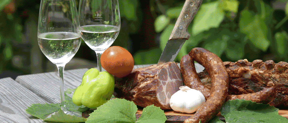 Zwei Weingläser mit Wasser, ein Messer und verschiedene Lebensmittel wie Tomate, Pfeffer und Wurst auf einem Tisch mit Blättern.
