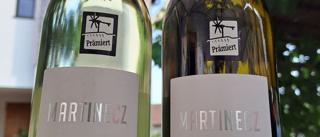 Zwei Weinflaschen von Martincz stehen nebeneinander. Eine Flasche ist mit 'Welschriesling 2024' und die andere mit 'Gelber Muskateller 2024' beschriftet. Beide Flaschen haben schwarze Etiketten mit dem Logo des Weinguts.