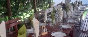 Ein langer roter Tisch ist mit weißen Tellern, Weingläsern und grünen Servietten bestückt. Er ist von roten Bänken und Holzpfosten umgeben. Der Hintergrund zeigt eine grüne Pflanze und ein Gebäude mit roten Regenschirmen.