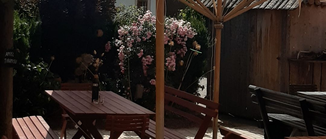 Ein Garten mit einer Holzbank und einem Tisch, bedeckt von einem beigen Regenschirm, mit einem Haus und Pflanzen im Hintergrund.