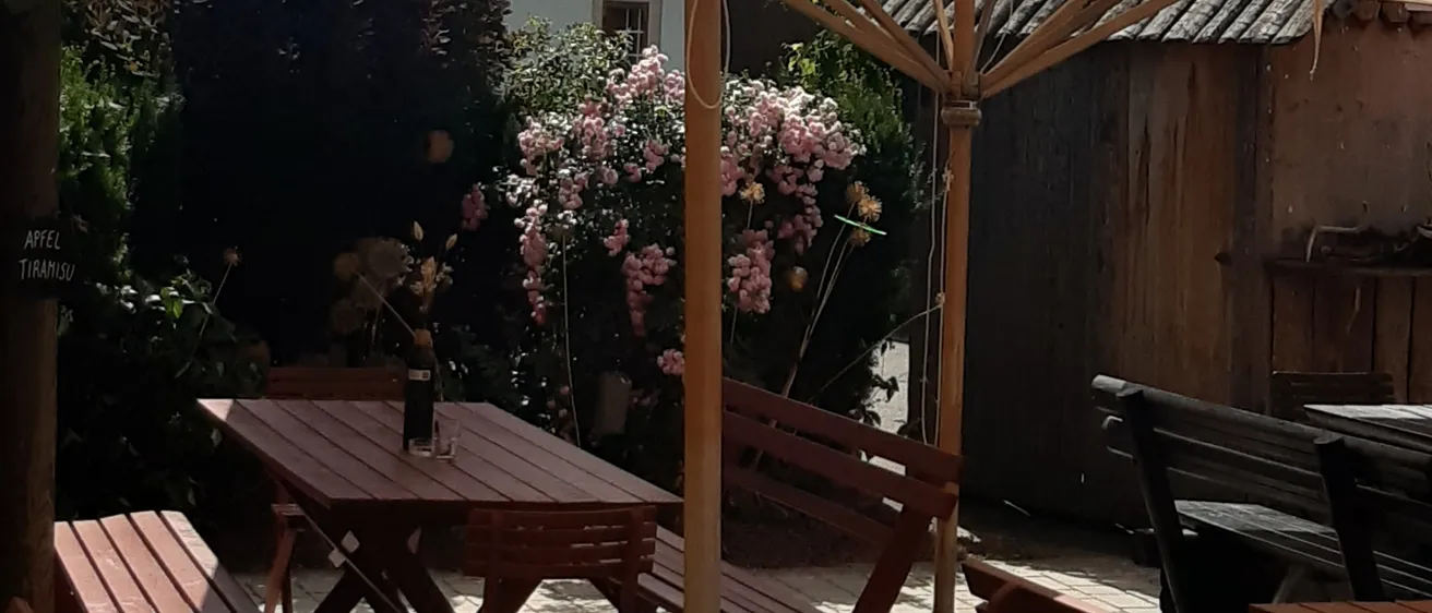 Ein Garten mit einer Holzbank und einem Tisch, bedeckt von einem beigen Regenschirm, mit einem Haus und Pflanzen im Hintergrund.