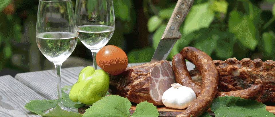 Ein Holzschneidebrett zeigt verschiedene Fleischsorten wie Salami, Prosciutto und Knoblauch. Zwei mit Wasser gefüllte Weingläser und eine grüne Paprika stehen daneben.