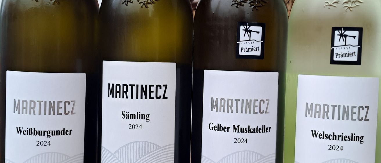 Vier Flaschen Wein stehen auf einem Holztisch. Jede Flasche hat ein Etikett mit 'Martinec' und dem Jahr 2024. Die erste Flasche ist mit 'Weiburgunder' beschriftet, die zweite mit 'Saumling', die dritte mit 'Gelber Muskateller' und die vierte mit 'Welschriesling'.