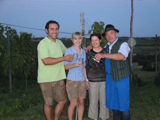 Vier Personen stehen in einem Weinberg. Zwei Männer und zwei Frauen. Ein Mann öffnet eine Flasche Wein, während die anderen Gläser halten.