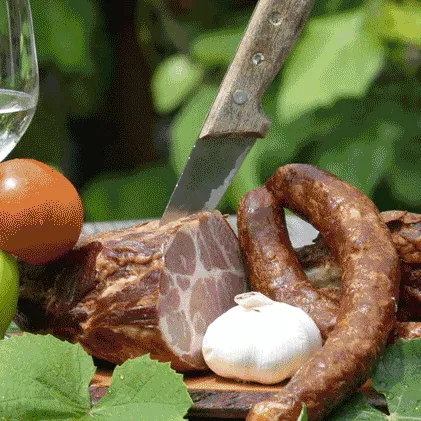 Ein Tisch mit zwei Weingläsern, einem Messer und einer Platte mit Fleisch, darunter Salami, Wurst und gepökeltes Fleisch. Eine grüne Paprika, eine Tomate und Knoblauch sind ebenfalls vorhanden.