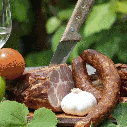 Ein Tisch mit zwei Weingläsern, einem Messer und einer Platte mit Fleisch, darunter Salami, Wurst und gepökeltes Fleisch. Eine grüne Paprika, eine Tomate und Knoblauch sind ebenfalls vorhanden.