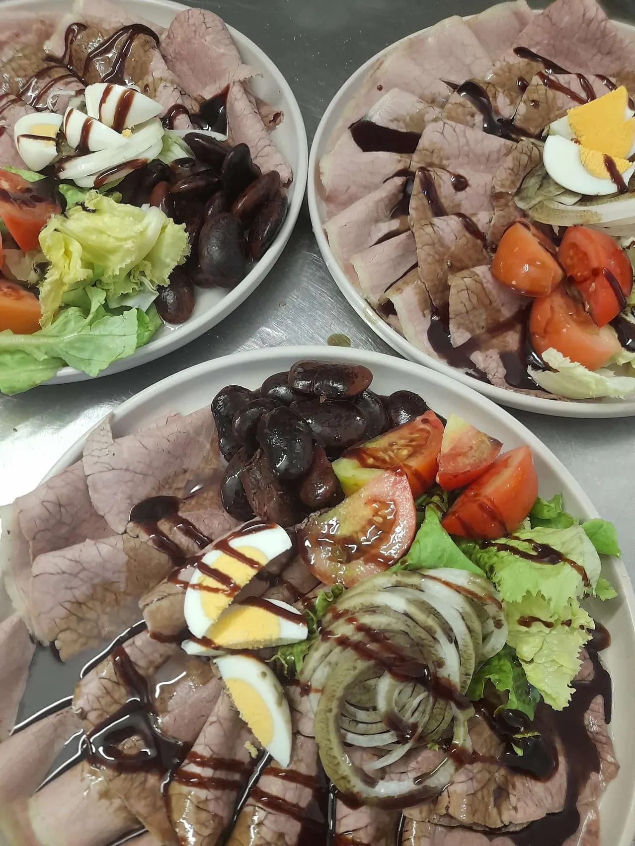 Drei Teller mit einer Mahlzeit aus Fleisch, Eiern und Gemüse. Jeder Teller hat eine Mischung aus geschnittenem Fleisch, Eiern und Gemüse, einschließlich Tomaten, Zwiebeln und schwarzen Bohnen.