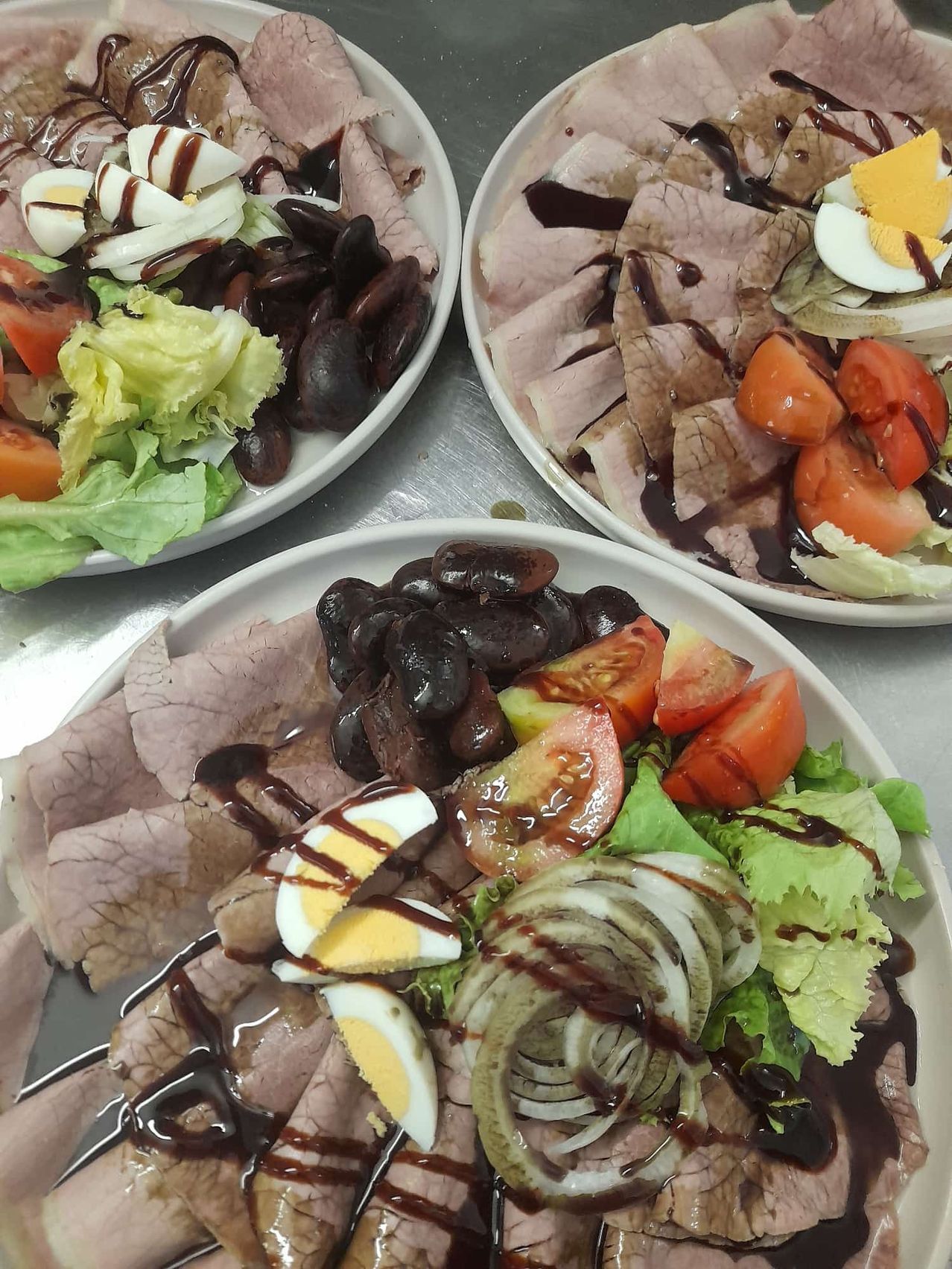 Drei Teller mit einer Mahlzeit aus Fleisch, Eiern und Gemüse. Jeder Teller hat eine Mischung aus geschnittenem Fleisch, Eiern und Gemüse, einschließlich Tomaten, Zwiebeln und schwarzen Bohnen.
