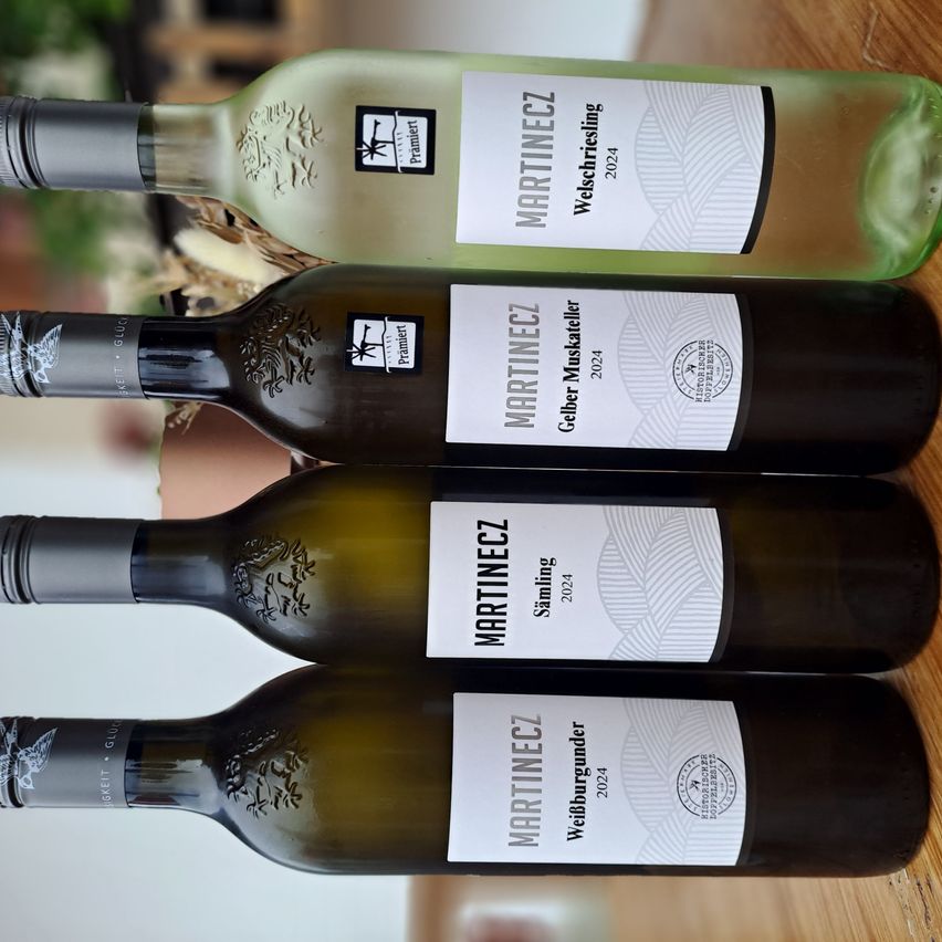 Vier Flaschen Wein von Martinecz sind gestapelt. Jede Flasche hat ein anderes Etikett: Welschriesling, Geiler Muskateller, Sämling und Weinbergunder. Die Flaschen haben einen klaren Glaskörper und dunkle Kappen.
