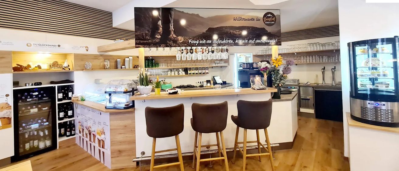 A cafe interior with three brown stools in front of a wooden counter, a coffee machine, and a plant on the right. A banner with the text 'Willkommen im Berghutt' is hanging above.
