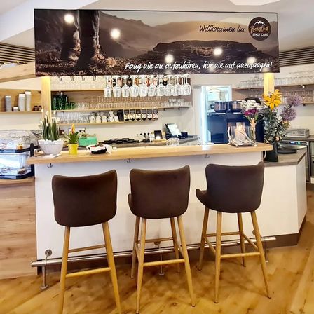 A cafe with a wooden counter, three stools, and a banner. The banner reads, 'Feel like you're listening, hear us out!' There are flowers, plants, and bottles on the counter.
