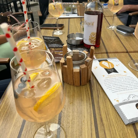 A wooden table displays a glass filled with a drink featuring blueberries, lemon, and ice. A bottle of alcohol is nearby. A menu and a book are also on the table.