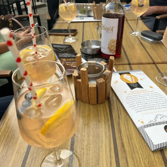 A wooden table displays a glass filled with a drink featuring blueberries, lemon, and ice. A bottle of alcohol is nearby. A menu and a book are also on the table.