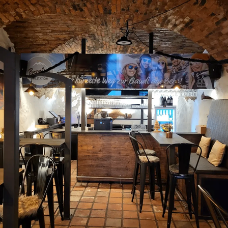 An interior of a cozy restaurant with brick walls and arched ceilings. A large banner reads 'kurzeste weg zur Gaudi am Berg!' A bar counter is equipped with stools, a bench, and shelves with bottles.