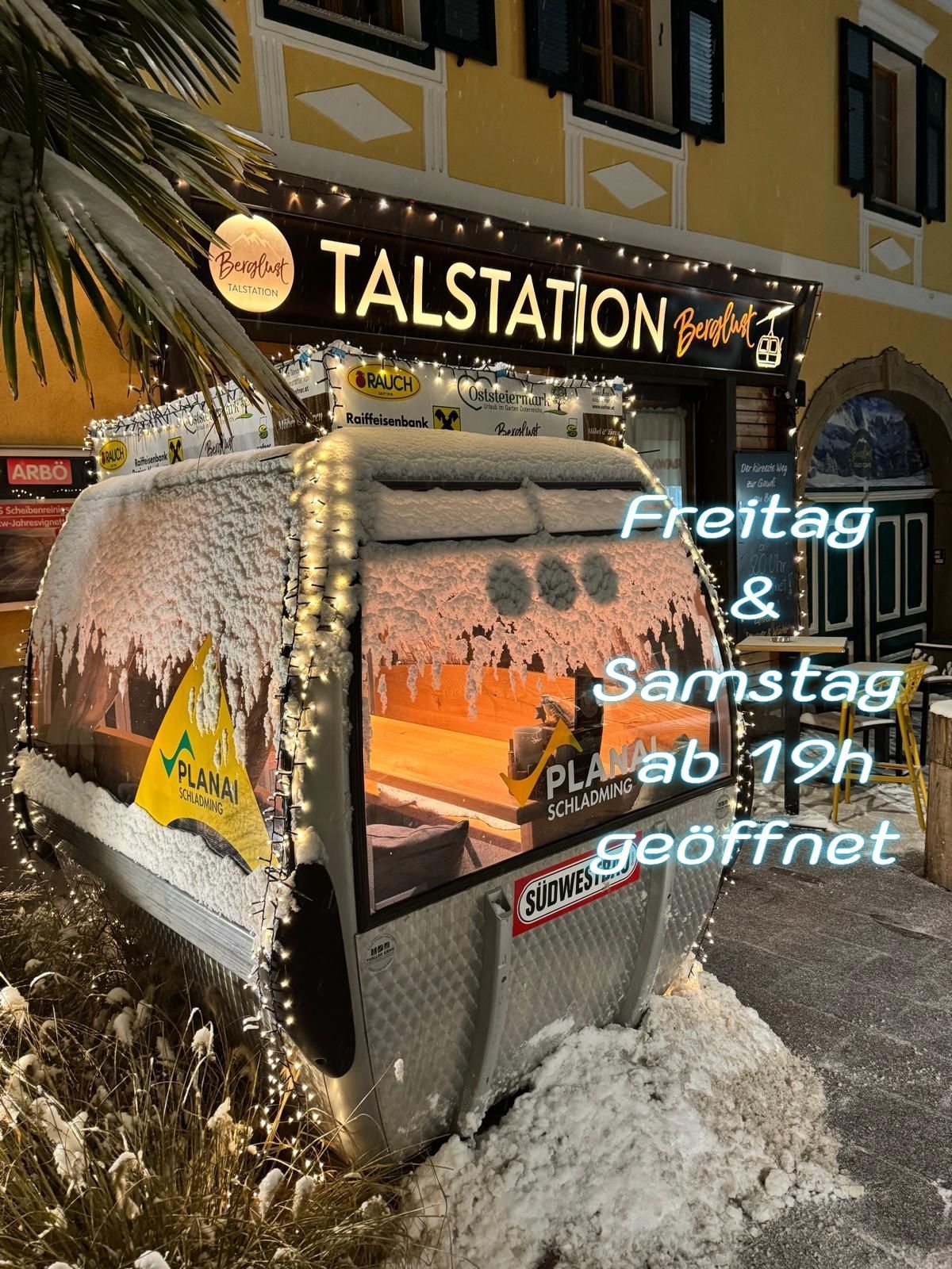 An outdoor area featuring a covered cable car with a sign that reads Planai Schladming. The signboard of the Talstation Bergfürst is covered with snow and Christmas lights. The text Freitag und Samstag ab 19h geoffnet is written on the ground.