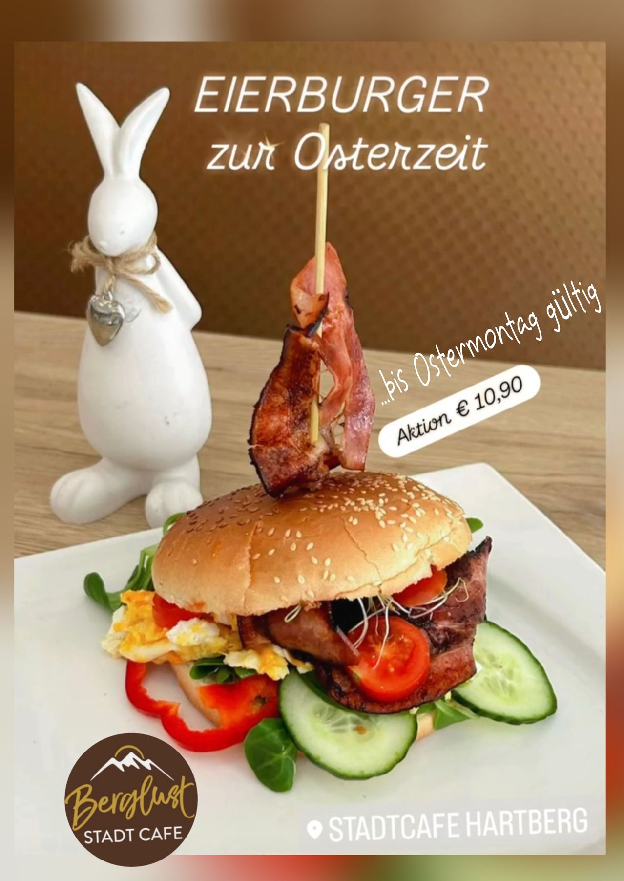 Ein Burger mit Speck, Eiern, Tomaten und Gurken auf einem Holztisch. Eine weiße Kaninchenfigur ist in der Nähe. Der Preis beträgt 10,90. Gültig bis Ostermontag.
