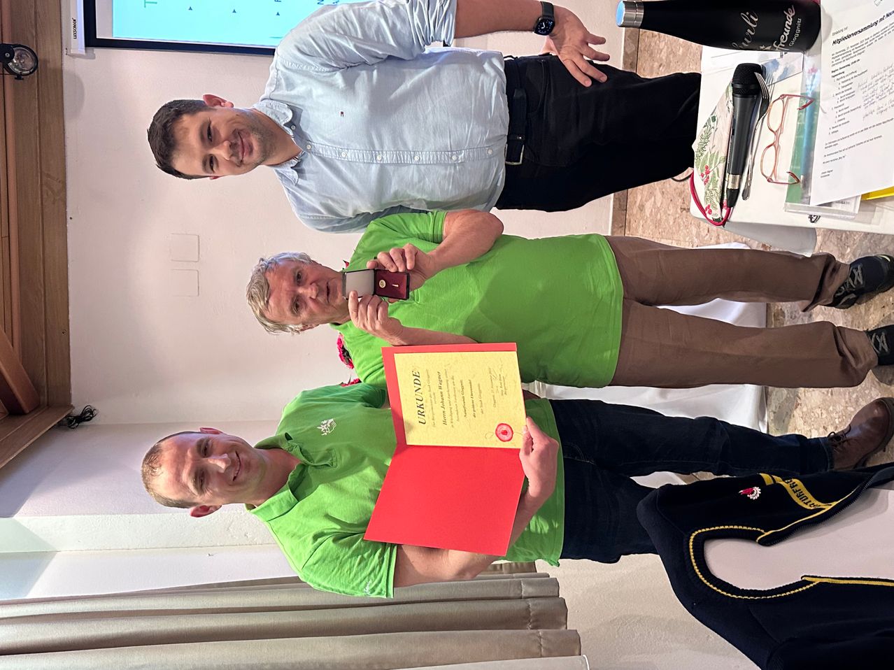 Drei Männer stehen in einem Raum, einer hält ein Zertifikat und lächelt. Ein Mann macht ein Foto mit einem Handy, und ein anderer hält ein Mikrofon.