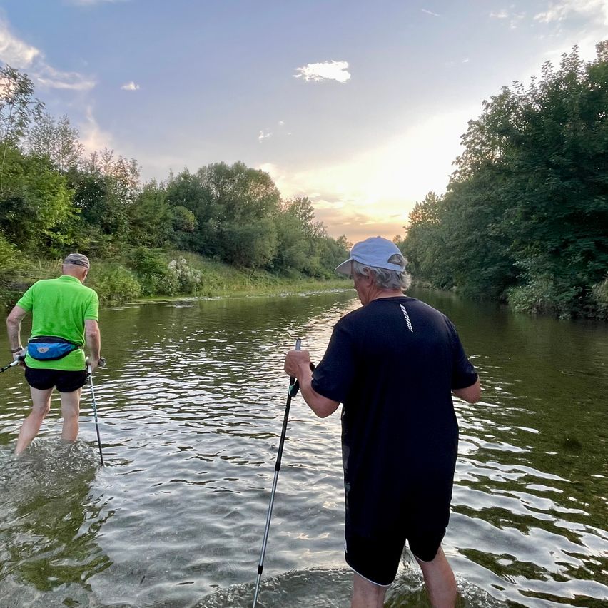Zwei Männer überqueren einen Fluss mit Trekkingstöcken, einer in einem grünen Shirt und der andere in einem schwarzen Shirt. Der Fluss ist von Bäumen und Büschen umgeben.