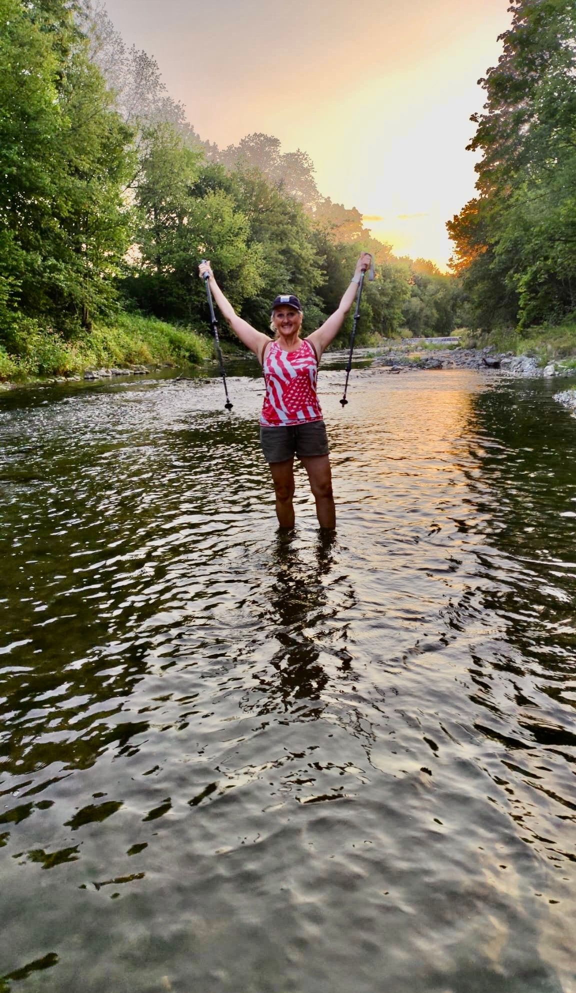 Eine Frau steht mit Wanderstöcken in einem Fluss, lächelt und ist von üppiger Vegetation und einer untergehenden Sonne umgeben.