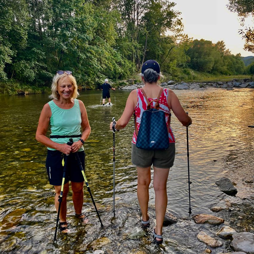 Drei Frauen stehen mit Wanderstöcken in einem Fluss, eine mit einem Rucksack. Hinter ihnen steht ein Mann im Wasser. Der Fluss ist von Bäumen umgeben.