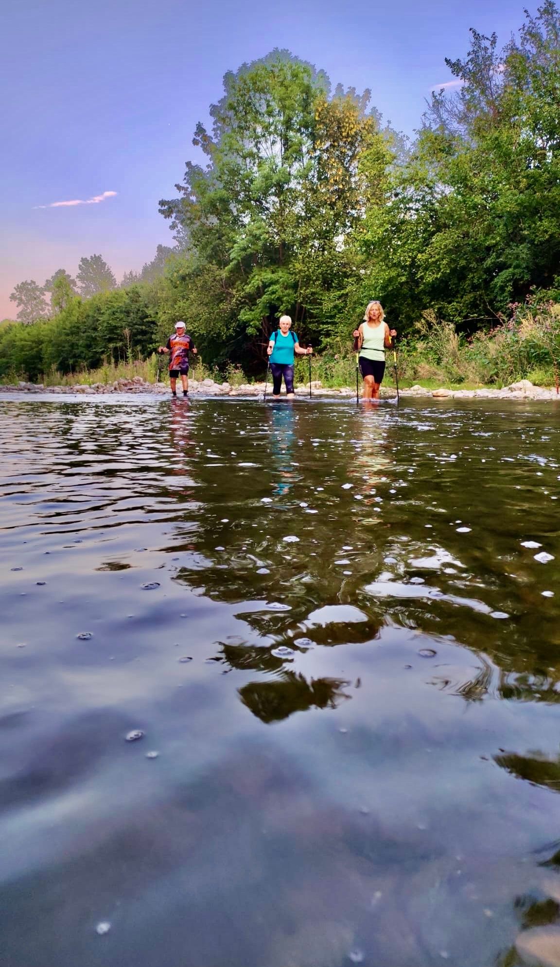 Drei Personen überqueren einen Fluss mit Trekkingstöcken. Der Fluss ist ruhig mit Wellen. Sie sind von Bäumen und Büschen umgeben. Der Himmel ist klar.