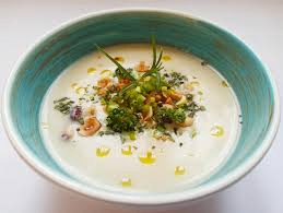 Eine weiße Schüssel mit einer cremigen Suppe, die mit Kräutern und Nüssen garniert ist. Die Suppe hat kleine Öltröpfchen, die auf ihrer Oberfläche schwimmen.