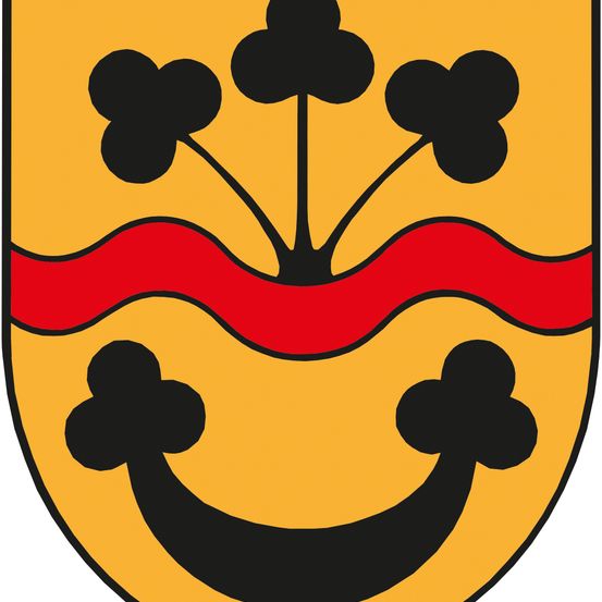 Das Emblem zeigt einen gelben Schild mit einer roten Wellenlinie und drei schwarzen Kleeblättern oben, die ein lächelndes Gesicht bilden.