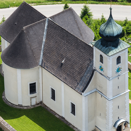 Ein Luftbild einer Kirche mit grünem Dach, weißen Mauern und einem hohen Turm. Der Turm hat Uhren auf zwei Seiten. Grüner Rasen umgibt das Gebäude.