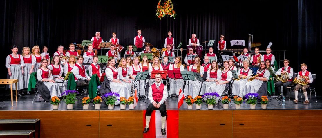 Eine große Gruppe von Menschen in traditioneller Kleidung posiert für ein Foto auf einer Bühne, mit Musikinstrumenten und Blumen im Hintergrund.
