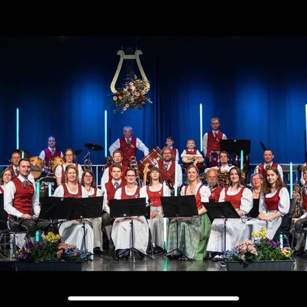 Ein Musikensemble in traditioneller Kleidung tritt auf der Bühne auf, mit Musikern, die Instrumente wie Trompeten, Trommeln und Flöten spielen. Die Bühne ist mit blauen Vorhängen und Blumen geschmückt.