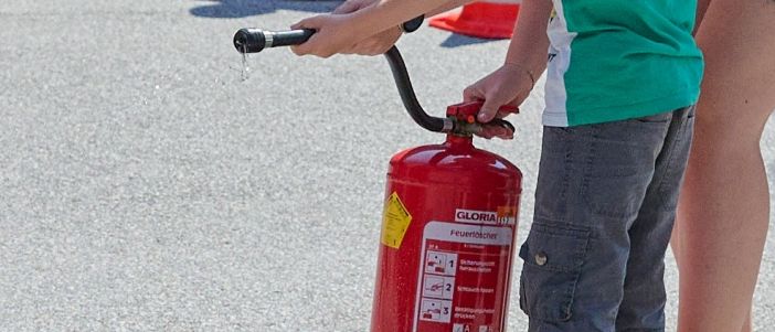 Ein junger Junge übt das Benutzen eines Feuerlöschers, während andere zusehen, einschließlich eines Jungen, der in der Nähe kniet. Die Szene spielt sich draußen auf einer Betonoberfläche ab.