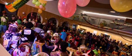 Eine lebhafte Indoor-Party mit Luftballons und Dekorationen. Menschen in Kostümen, Hüten und Masken, einige mit Plakaten, unterhalten sich. Die Umgebung ist festlich, mit Lichtern und einer lebhaften Atmosphäre.