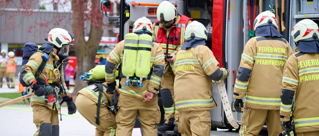 Eine Gruppe Feuerwehrleute in Uniform versammelt sich um einen Feuerwehrwagen und bereitet sich auf einen Einsatz vor. Einer hält eine Feuerlöscher.