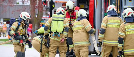 Eine Gruppe Feuerwehrleute in Uniform versammelt sich um einen Feuerwehrwagen und bereitet sich auf einen Einsatz vor. Einer hält eine Feuerlöscher.