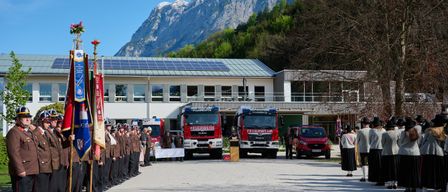 Eine Feuerwehr-Zeremonie mit zwei Feuerwehrwagen vor einem Gebäude mit Solarpaneelen und Fahnen. Die Berge sind im Hintergrund.