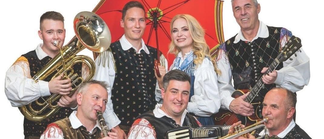 Eine Gruppe von Musikern in traditioneller Kleidung, die mit verschiedenen Instrumenten wie Trompete, Tuba, Klarinette und Akkordeon spielen. Sie stehen und sitzen mit einem Regenschirm im Hintergrund.
