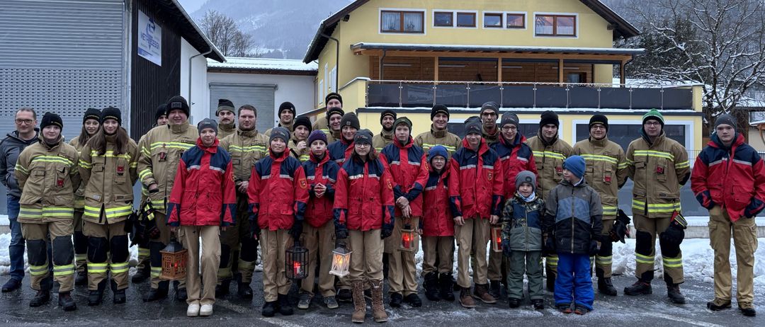 Eine Gruppe von Feuerwehrleuten, Erwachsene und Kinder, steht in Winteruniformen. Sie halten Laternen und posieren für ein Foto vor einem gelben Haus mit einem Berg im Hintergrund.