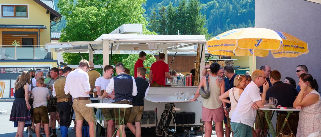 A group of people are gathered around a food truck. Some are standing, others are seated. The food truck is under a yellow umbrella. The background shows mountains and trees.