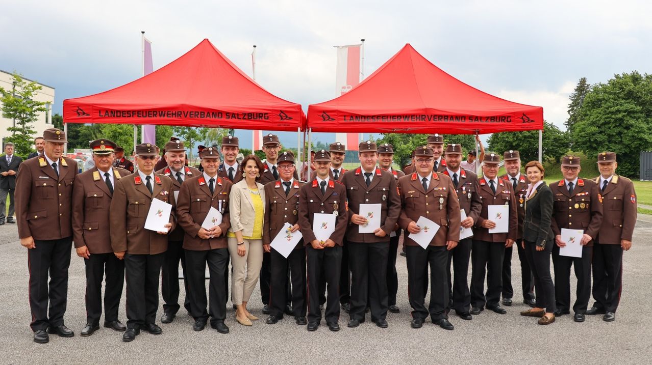 Eine Gruppe Feuerwehrleute in Uniform posiert für ein Foto, während sie Urkunden halten, mit zwei roten Zelten und Fahnen im Hintergrund.