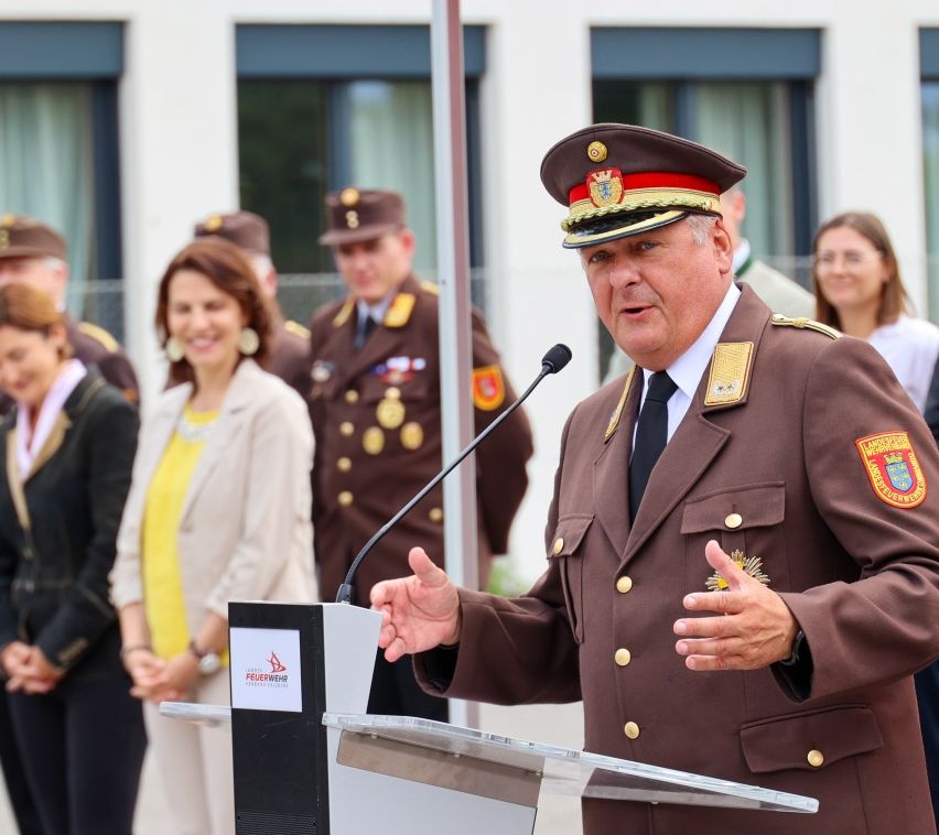 Ein Militärbeamter in Uniform spricht an einem Rednerpult mit Mikrofon, umgeben von anderen uniformierten Personen.