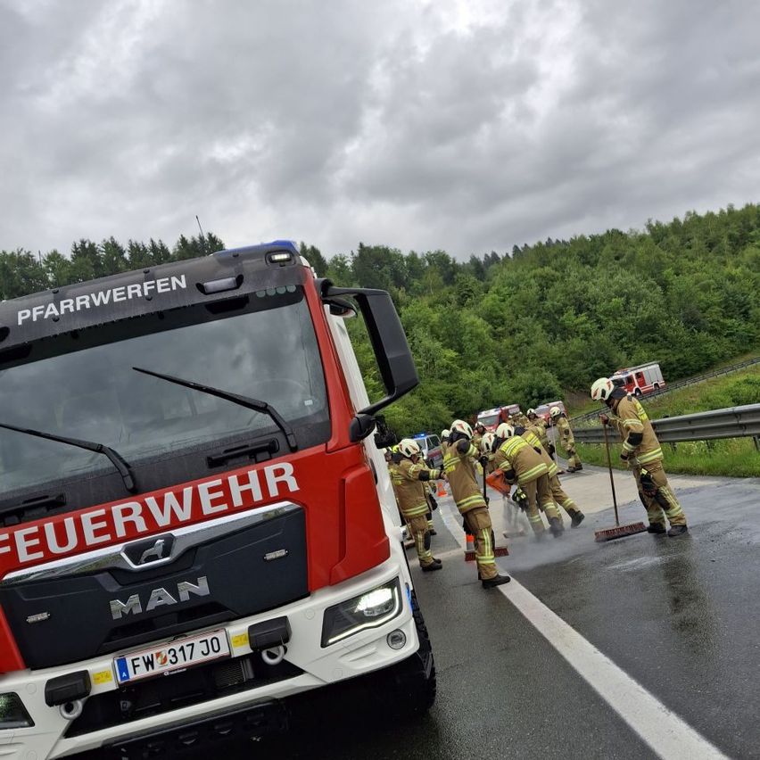 Eine Gruppe von Feuerwehrleuten reinigt die Straße vor einem roten Feuerwehrauto mit dem Wort 'Feuerwehr' auf der Front. Die Feuerwehrleute tragen Helme und Uniformen.