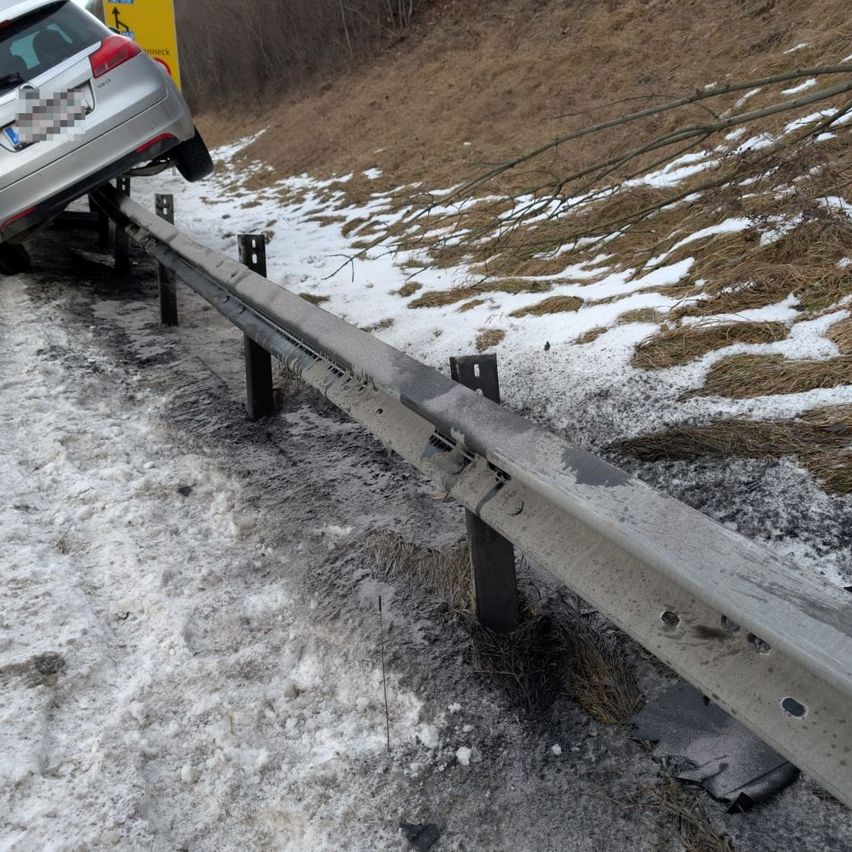 Ein silberner Wagen mit einem Nummernschild ruht auf einem Leitplanken. Das Auto hat seine Rückseite auf der Leitplanke. Es liegt Schnee auf dem Boden und ein Ast liegt auf dem Boden.