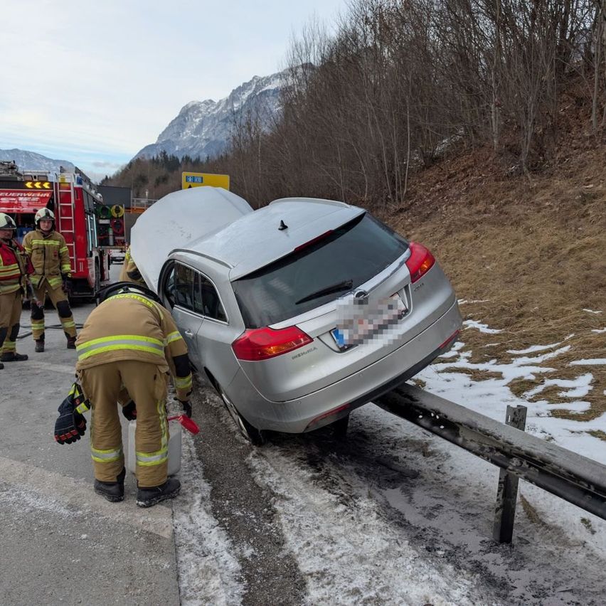 Feuerwehrleute kümmern sich um ein umgekipptes silbernes Auto auf einer verschneiten Straße. Zwei Feuerwehrleute sind in der Nähe, einer hält ein Werkzeug. Ein Feuerwehrauto ist im Hintergrund geparkt.