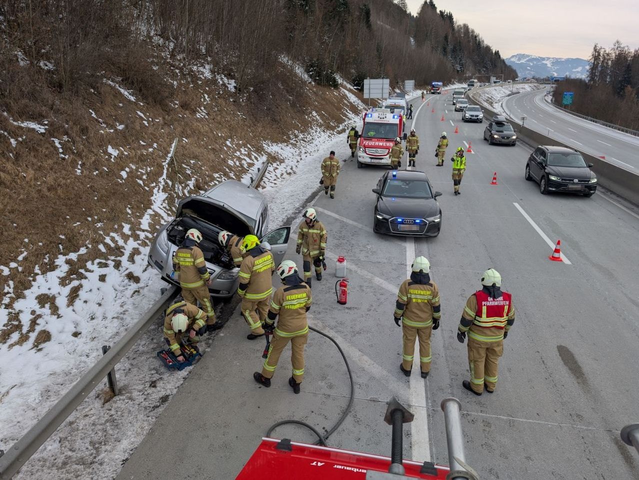Rettungskräfte in Schutzkleidung kümmern sich auf einer verschneiten Autobahn um ein umgekipptes Auto, mit Warnbaken und Feuerlöschern in der Nähe.