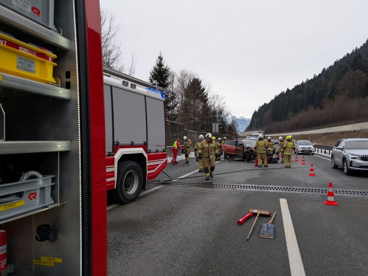 Ein Feuerwehrauto ist am Straßenrand geparkt, während Feuerwehrleute einen Autounfall bearbeiten. Die Szene zeigt ein beschädigtes Auto, Verkehrskegel und Schutzausrüstung.
