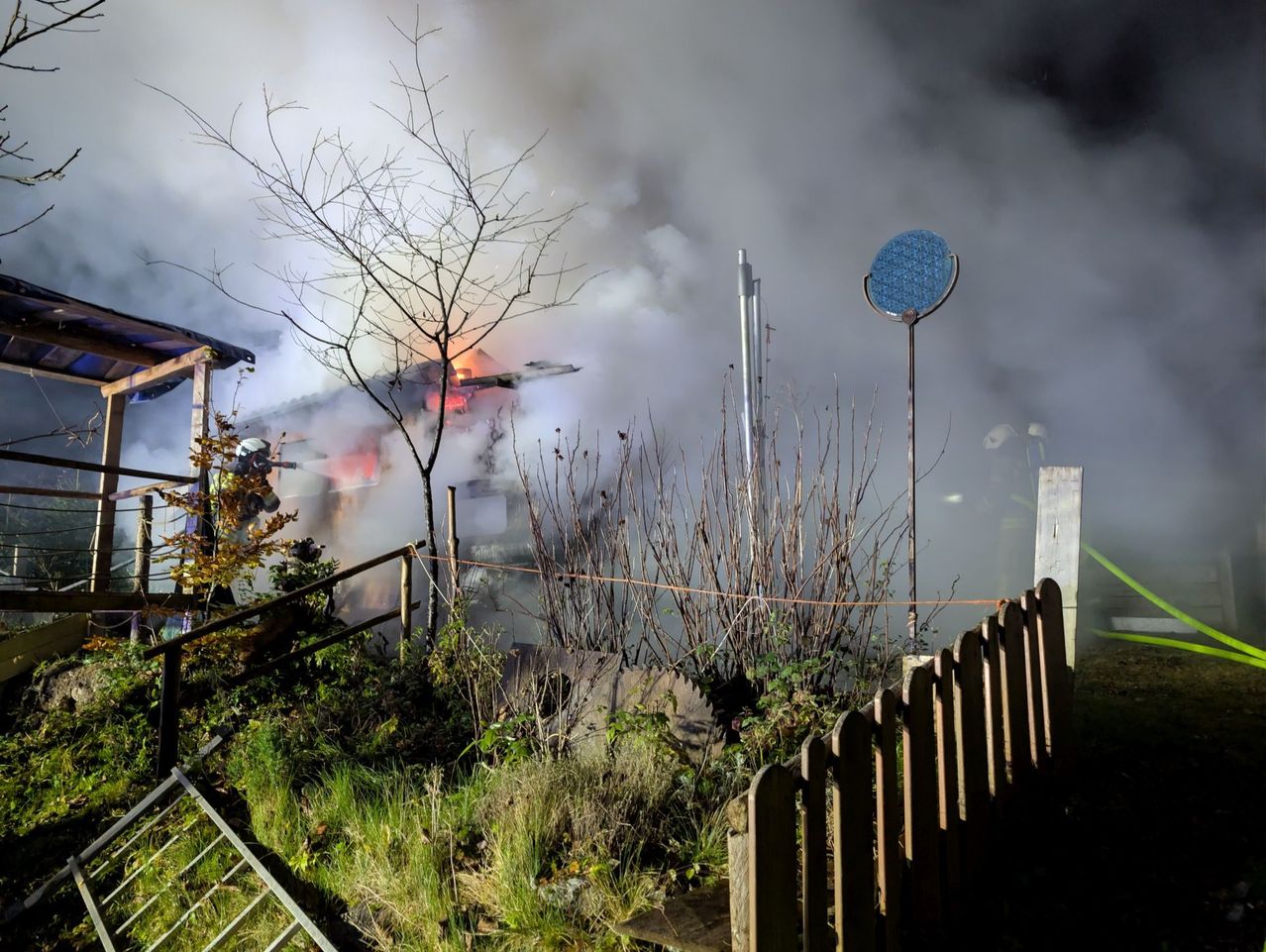 Firefighters are battling a blaze at a house, with smoke billowing from the roof. Nearby, a wooden fence and various structures are visible.