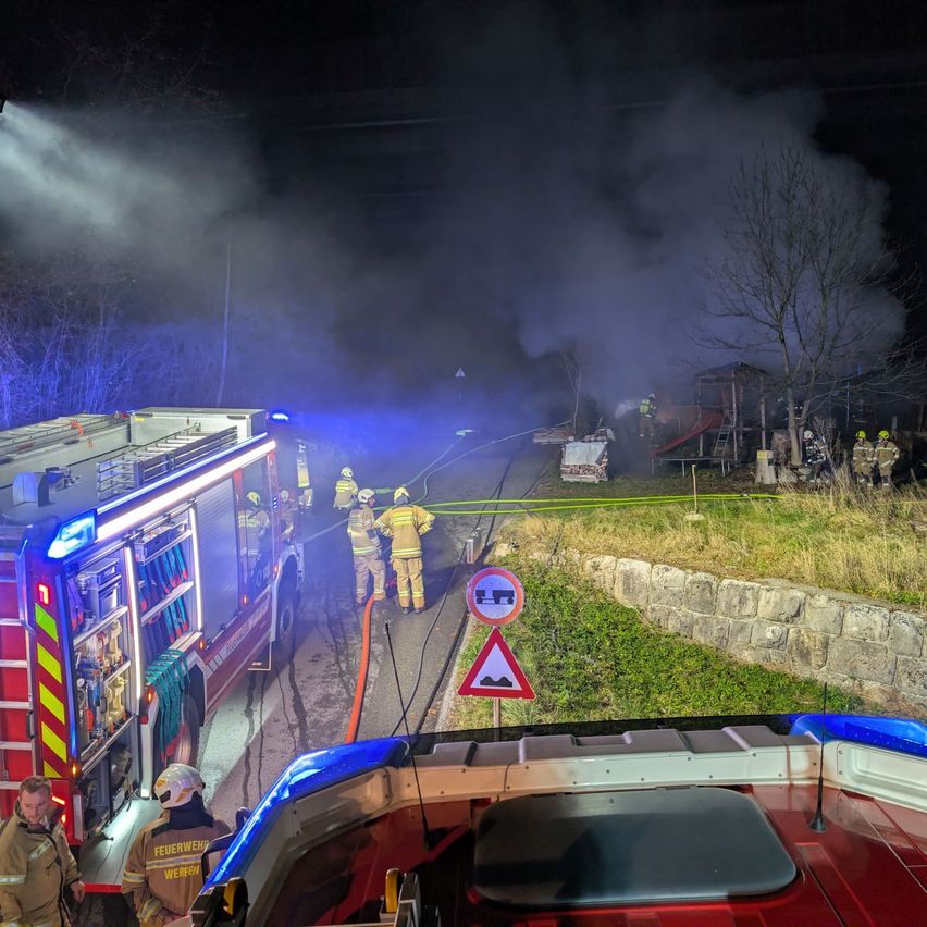 Firefighters are battling a blaze in a rural area at night. Several fire trucks and personnel are present, with smoke rising from a burning structure.