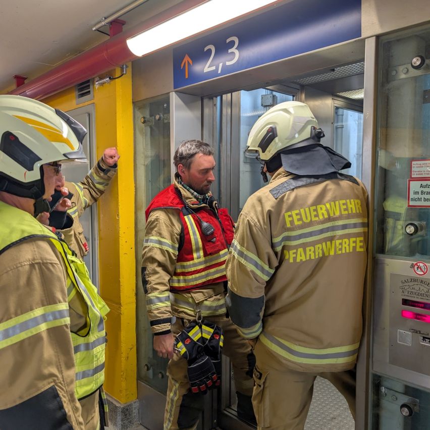 Drei Feuerwehrleute in voller Montur stehen vor einem Aufzug in einer U-Bahn-Station. Einer gestikuliert, während die anderen beiden zusehen.