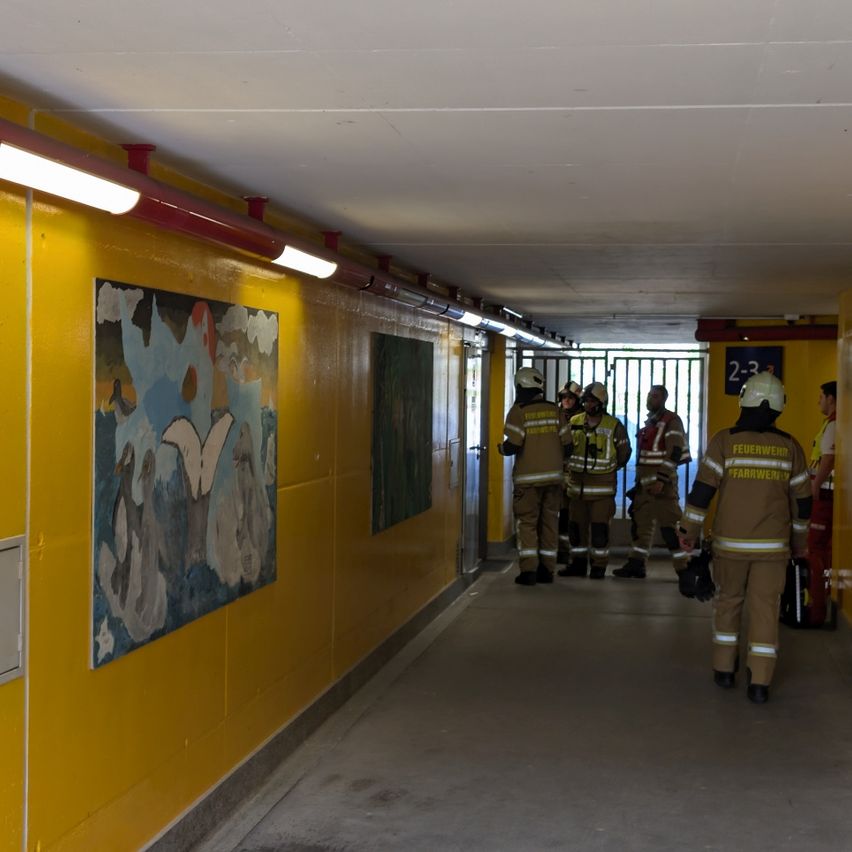 Feuerwehrleute in gelben Uniformen laufen in einem Tunnel mit gelben Wänden, beleuchtet von Deckenleuchten. Ein großes Gemälde ist an der Wand.